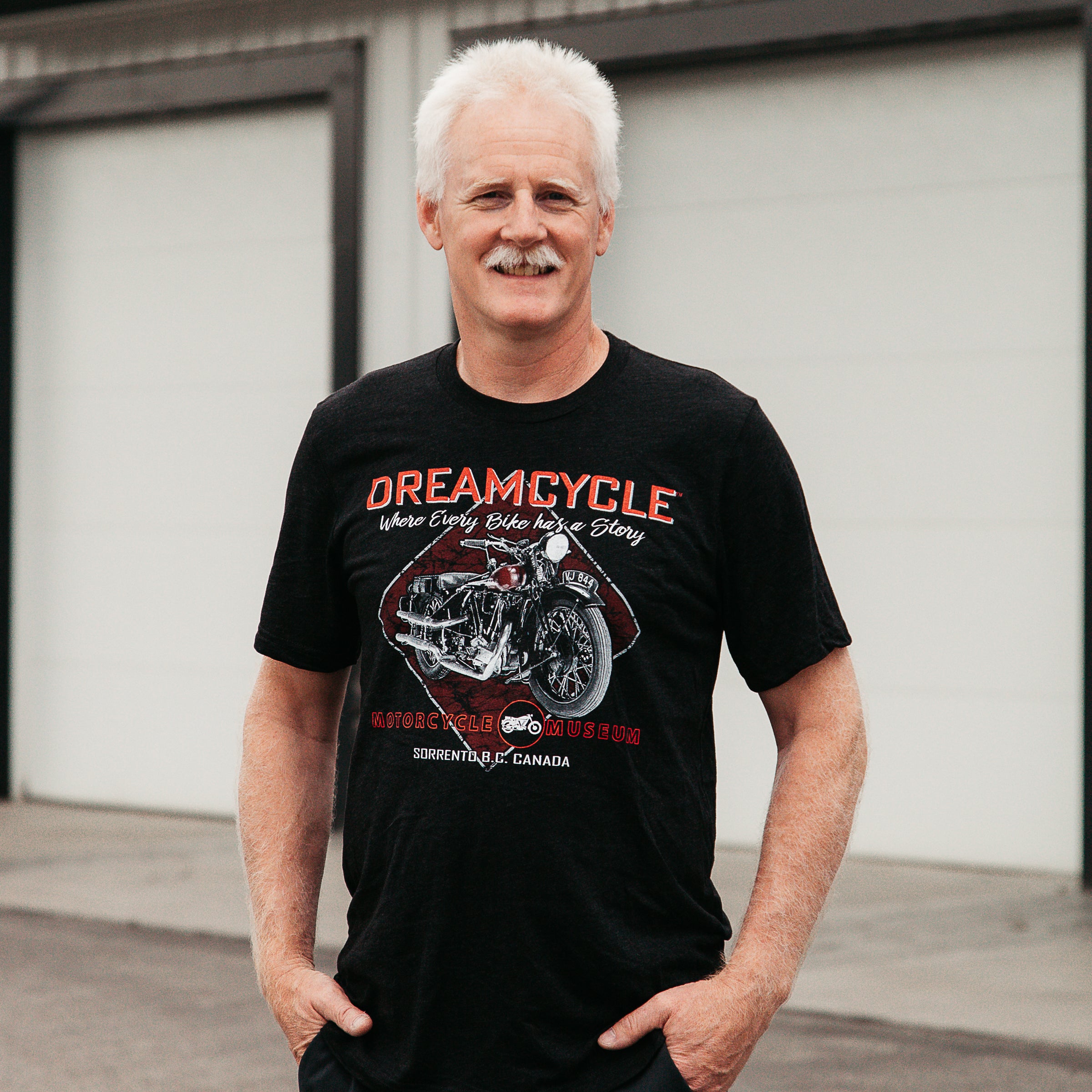 Dreamcycle Motorcycle Museum |  Man posing in lifestyle setting wearing a tshirt that says "Dreamcycle" in bold letters with a motorcycle.