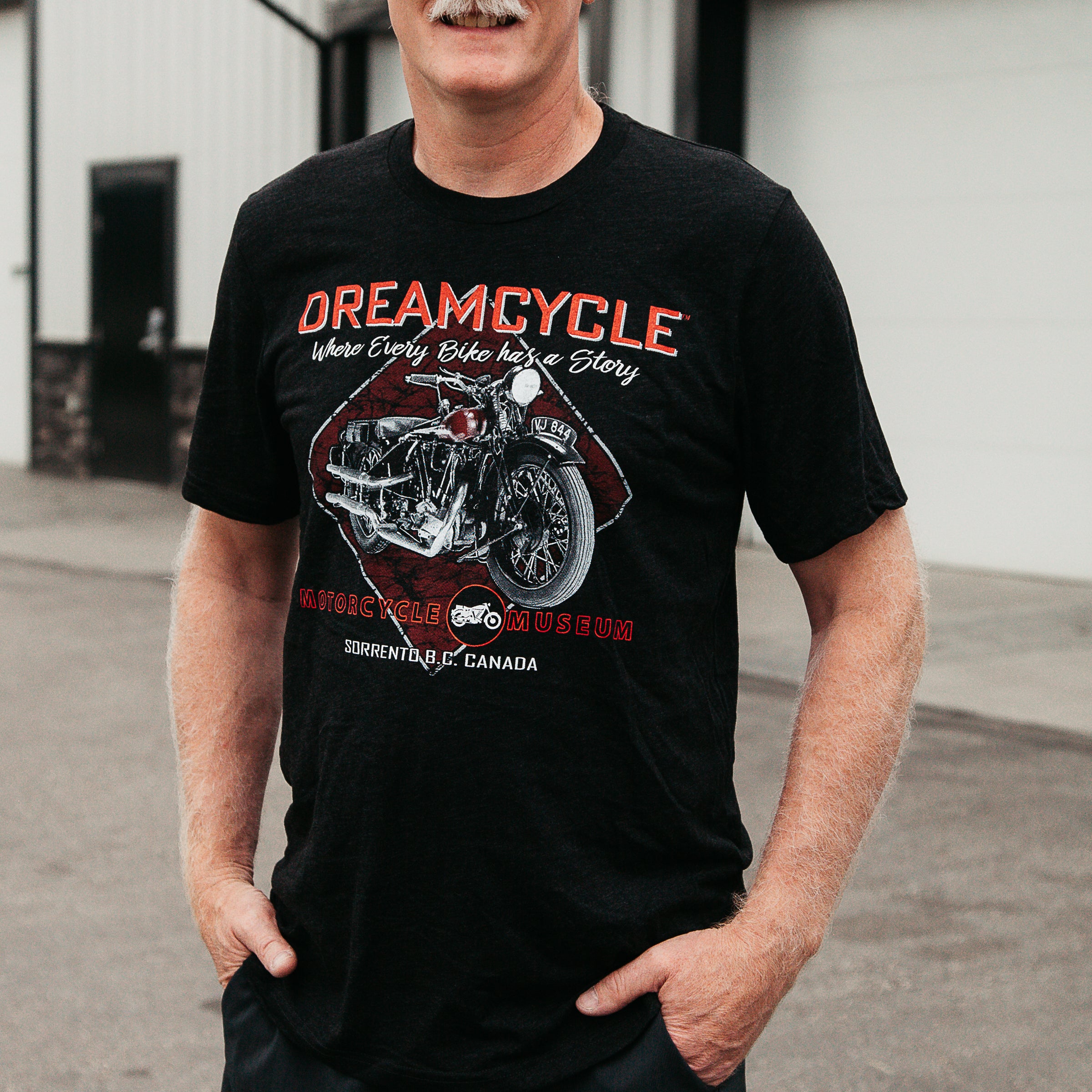 Dreamcycle Motorcycle Museum |  Close up of a man standing in a lifestyle setting wearing a shirt that says "`dreamcycle" With a motorcycle on the front.